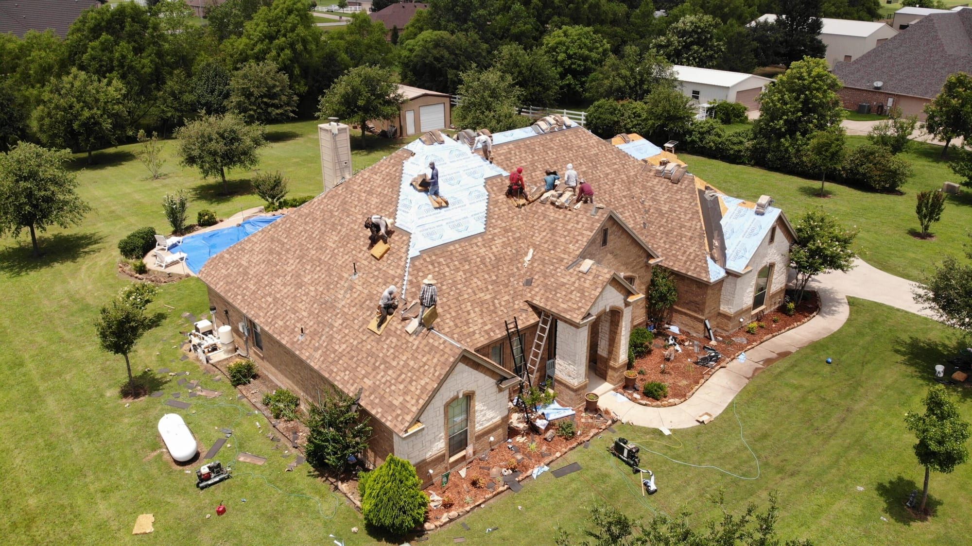 Crew starting the tear-off of an old brown shingle roof on a stone and stucco home, seen from a drone