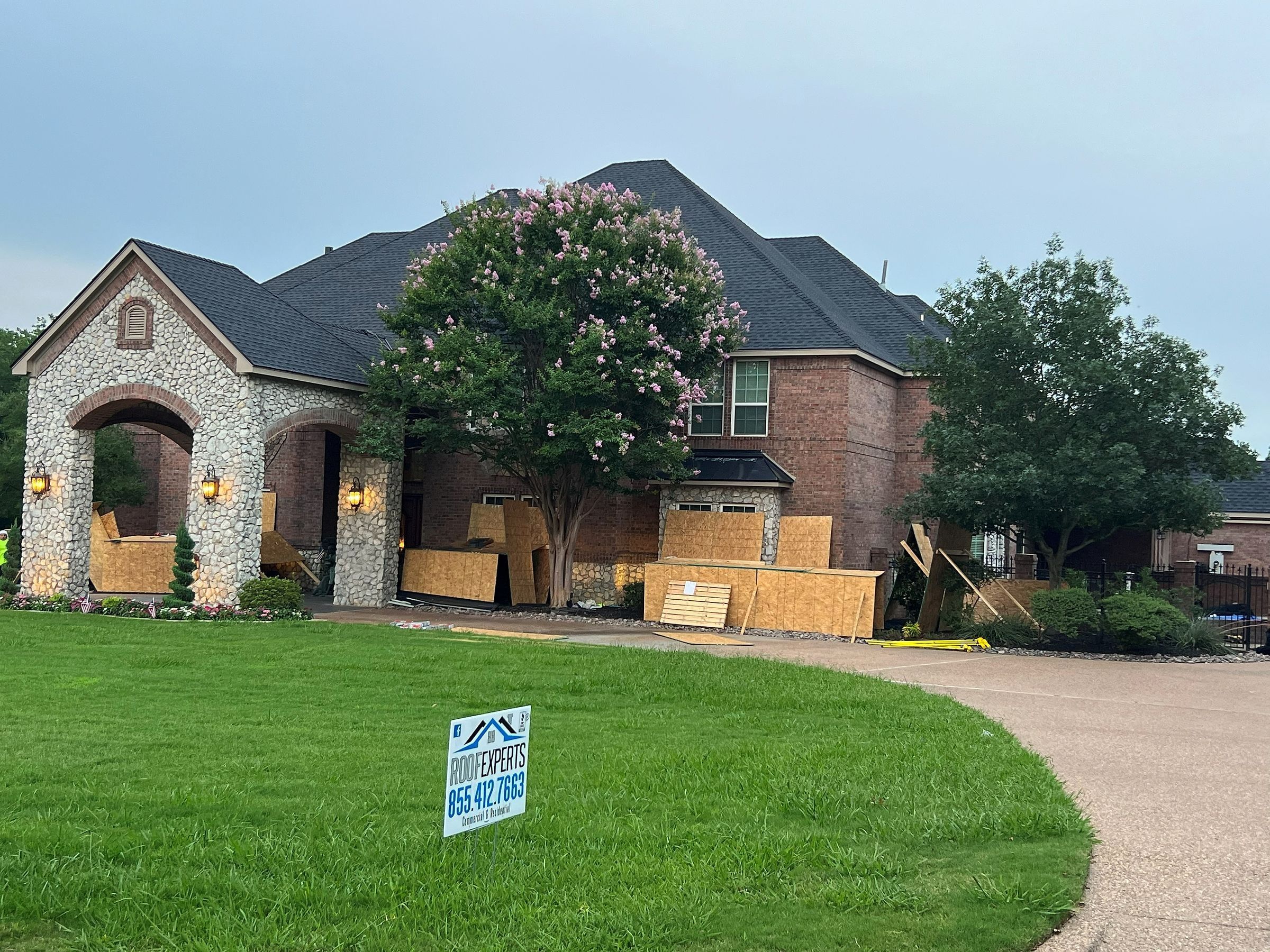 Plywood over the front windows of a brick home during a roof replacement, Roof Experts yard sign on the lawn