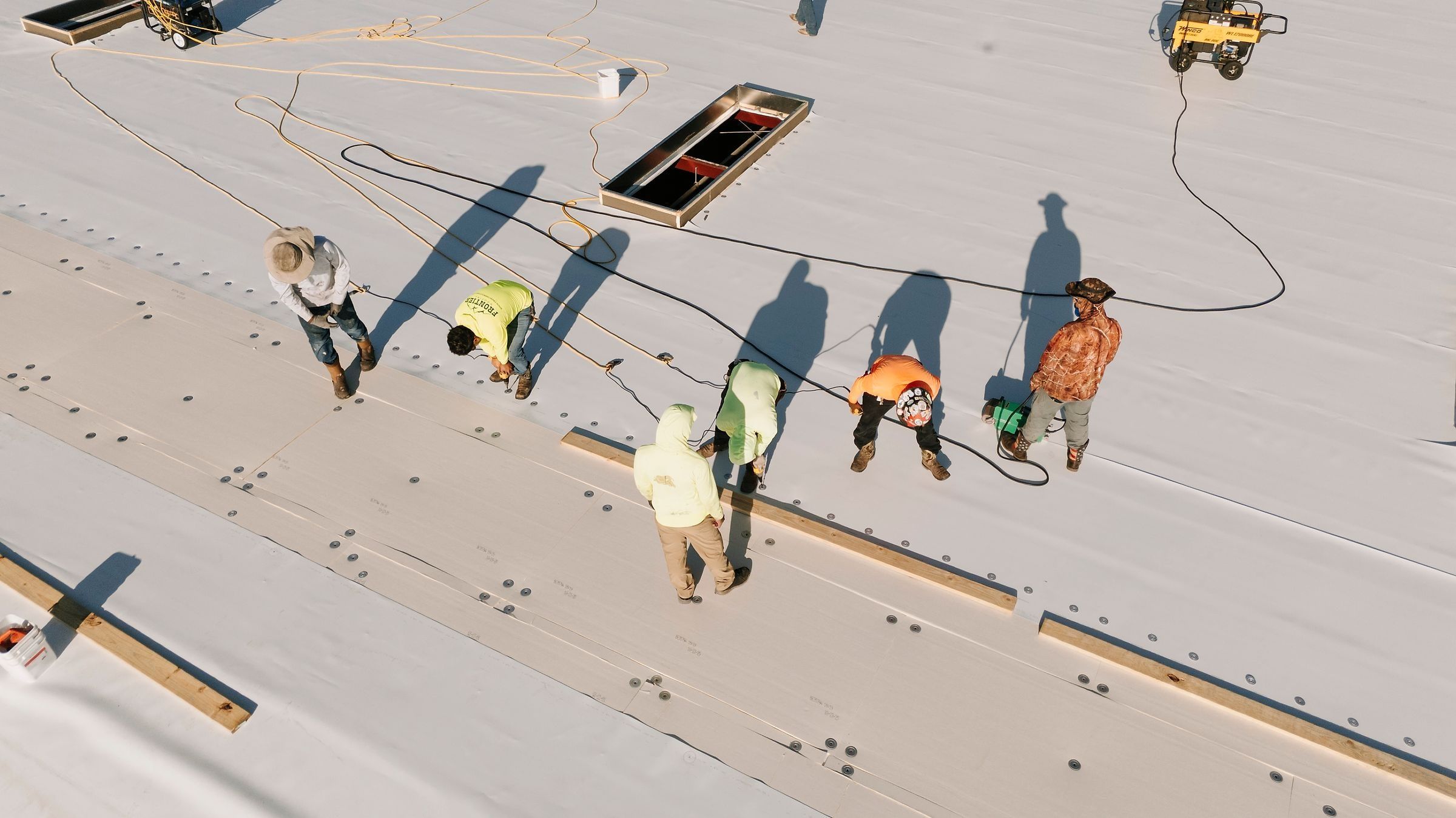 Six roofers on a white TPO roof with seam-welding equipment, seen from above